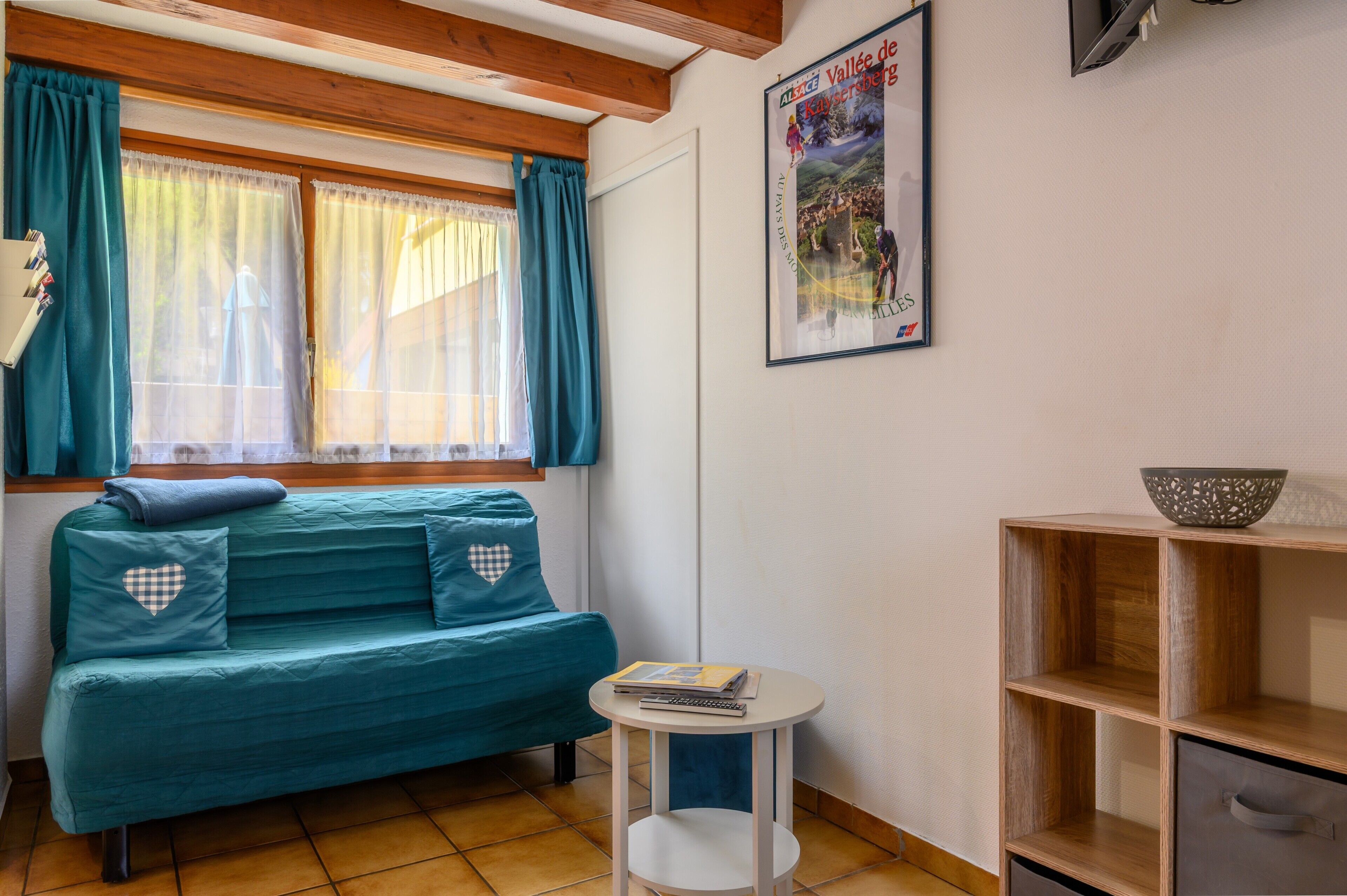 Um quarto com um sofá azul-turquesa, uma pequena mesa branca, uma estante de madeira e um cartaz na parede.