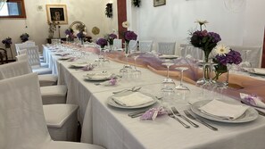 Een lange feesttafel gedekt met witte tafelkleden, glazen en paarse bloemen.