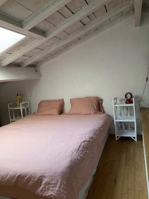 Une chambre avec un plafond en pente, un lit avec une literie rose, une table de chevet avec une lampe et une petite étagère sur laquelle se trouve un objet rouge.