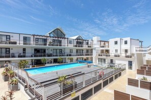 Un hotel moderno con piscina en la azotea, varios balcones y un cielo azul despejado.