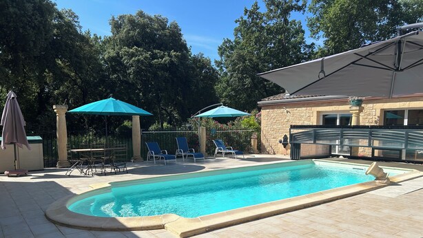 Une piscine entourée d’un mur de pierre et d’arbres, avec des chaises longues et des parasols à proximité.