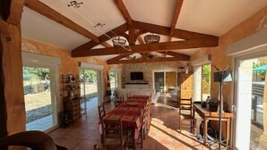 Une salle à manger spacieuse avec des poutres en bois, une longue table et des chaises. On y trouve également un coin cuisine avec un téléviseur et des placards en arrière-plan.