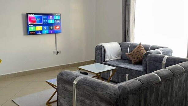 A modern living room with a grey sofa, a small coffee table, a flat-screen TV mounted on the wall, and a large window with curtains. - Snowhaven Kitengela| Modern, Secure Stay near Town (Kitengela)