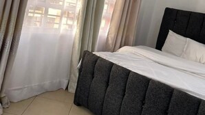 A bedroom with a bed, curtains, and a view of buildings through the window. - Snowhaven Kitengela| Modern, Secure Stay near Town (Kitengela)