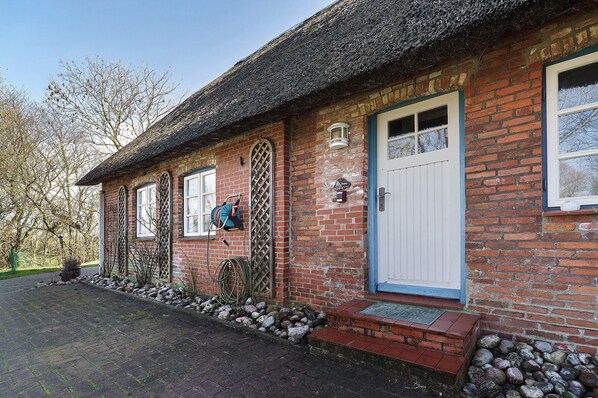 Ein Backsteinhaus mit weißer Tür, einem Fenster mit Gitterwerk und einem an der Wand aufgerollten Schlauch.