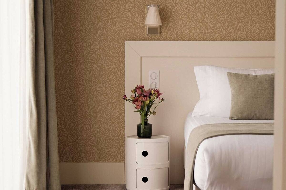 Een slaapkamer met een bed, nachtkastje, lamp en een vaas met bloemen.