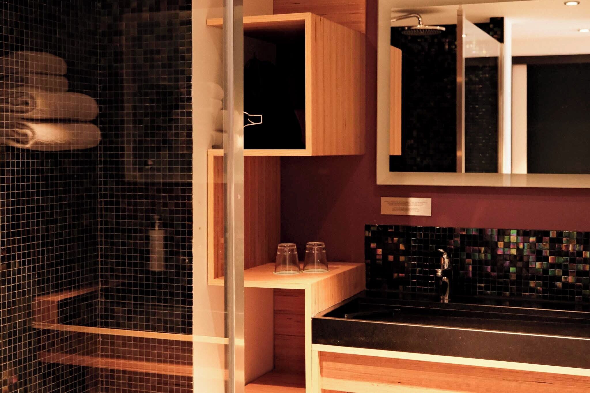 A modern bathroom with a glass shower enclosure, wooden cabinetry, and a black tiled backsplash.