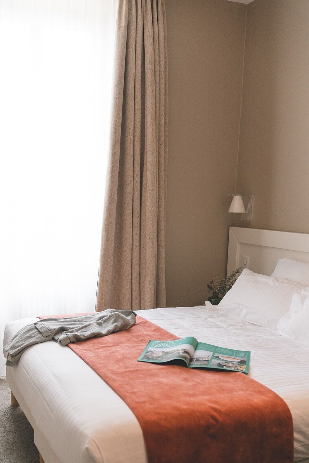 Une chambre d’hôtel avec un lit, une fenêtre avec des rideaux, une lampe et un magazine posé sur le lit.