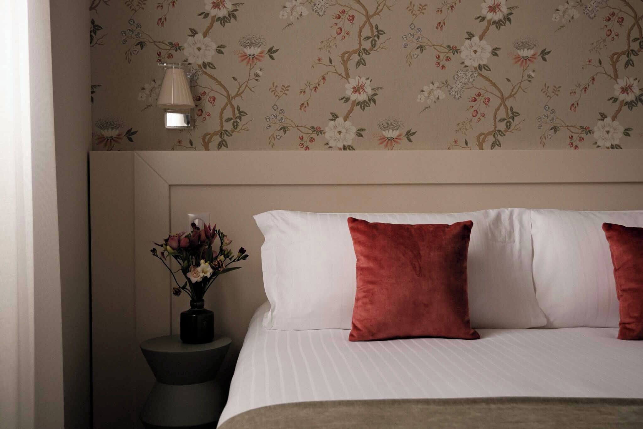 A neatly made bed with white and red pillows, a vase with flowers on a bedside table, and a floral wallpaper pattern.