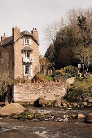 Un bâtiment en pierre doté d’un balcon, avec une rivière en contrebas et une végétation luxuriante tout autour.