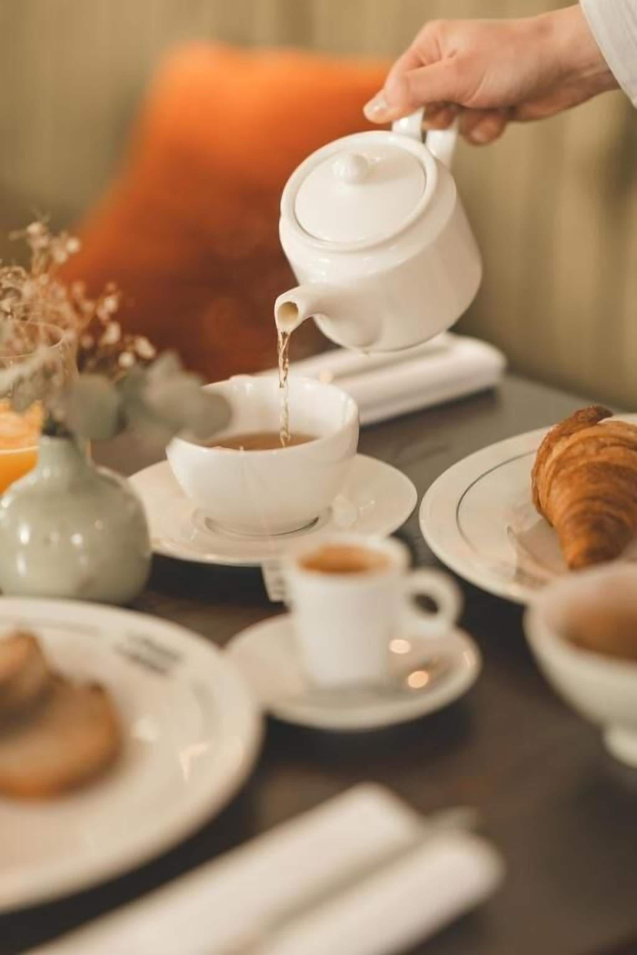 A table with a teapot pouring into a cup, a croissant, and a vase with dried flowers.