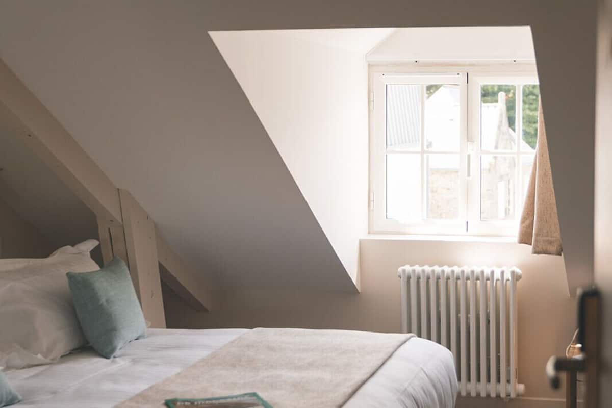 Een slaapkamer met een schuine kap, een raam met uitzicht, een radiator en een bed met kussens.