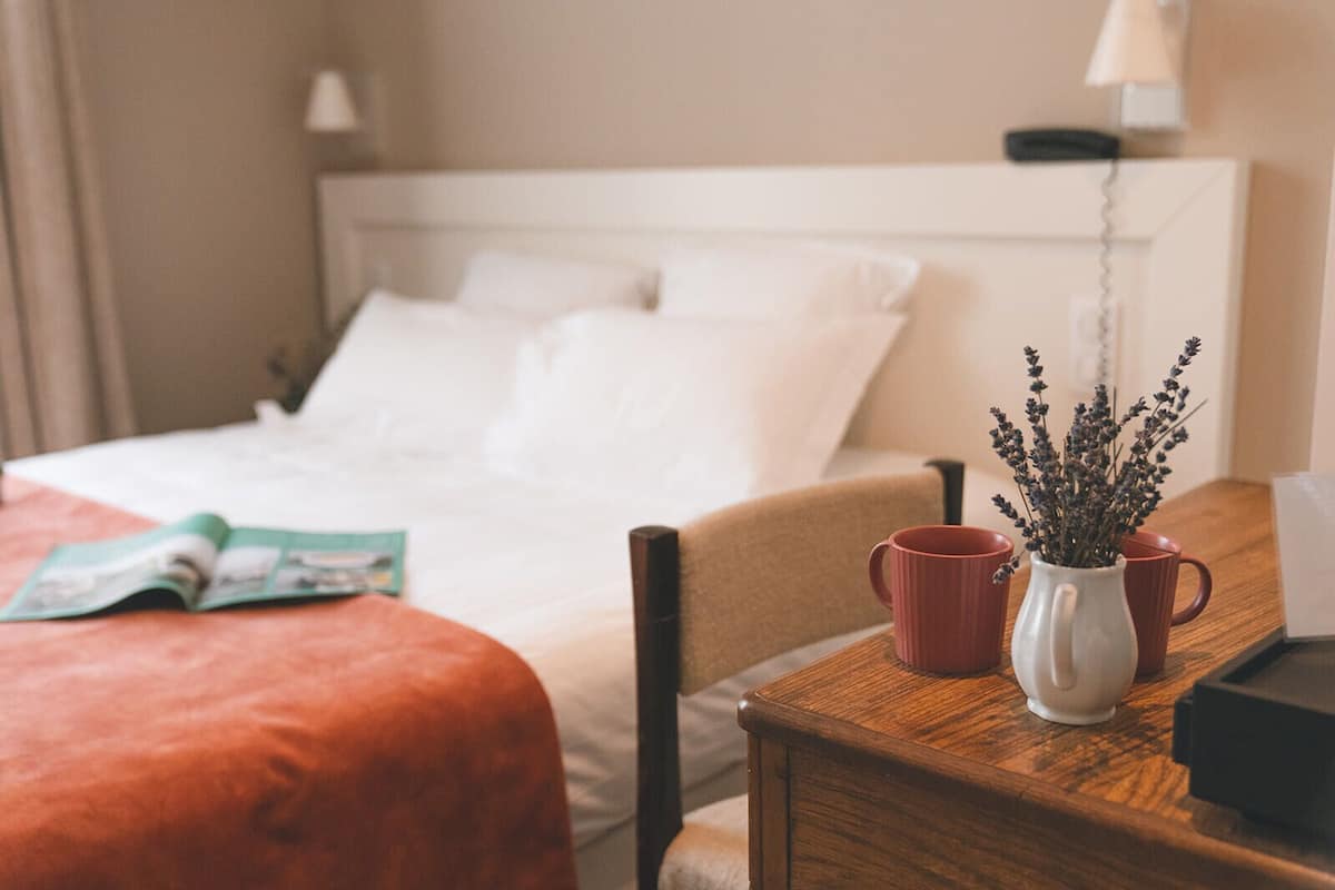Een hotelkamer met een bed, een houten nachtkastje, een rode mok en een vaas met gedroogde bloemen.