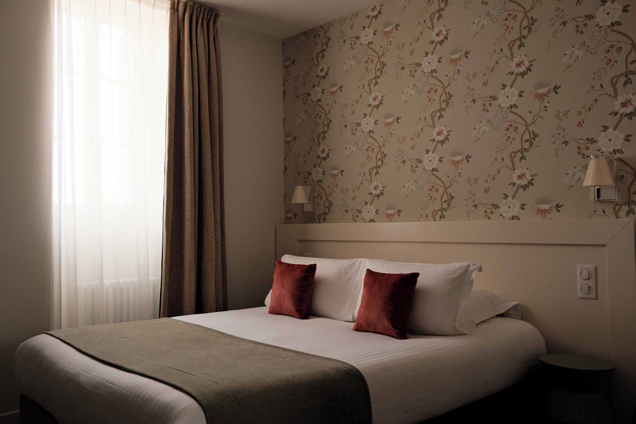 A hotel room with a bed, curtains, floral wallpaper, and a nightstand with a lamp.