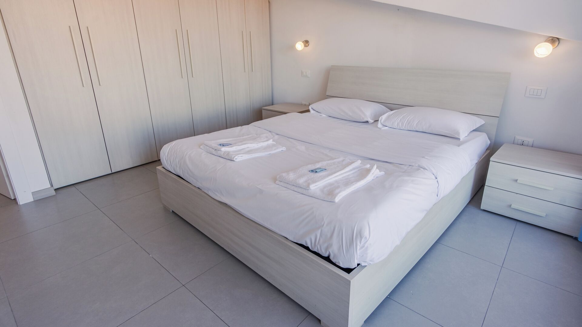 Une chambre moderne avec un grand lit, une table de chevet, une armoire et un luminaire fixé au mur.