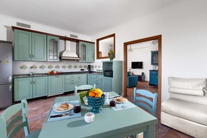 A kitchen with teal cabinets, a dining table with a fruit bowl, and a sofa in the adjacent living area. - Estate4home - Casa Marelia (Sorrento)