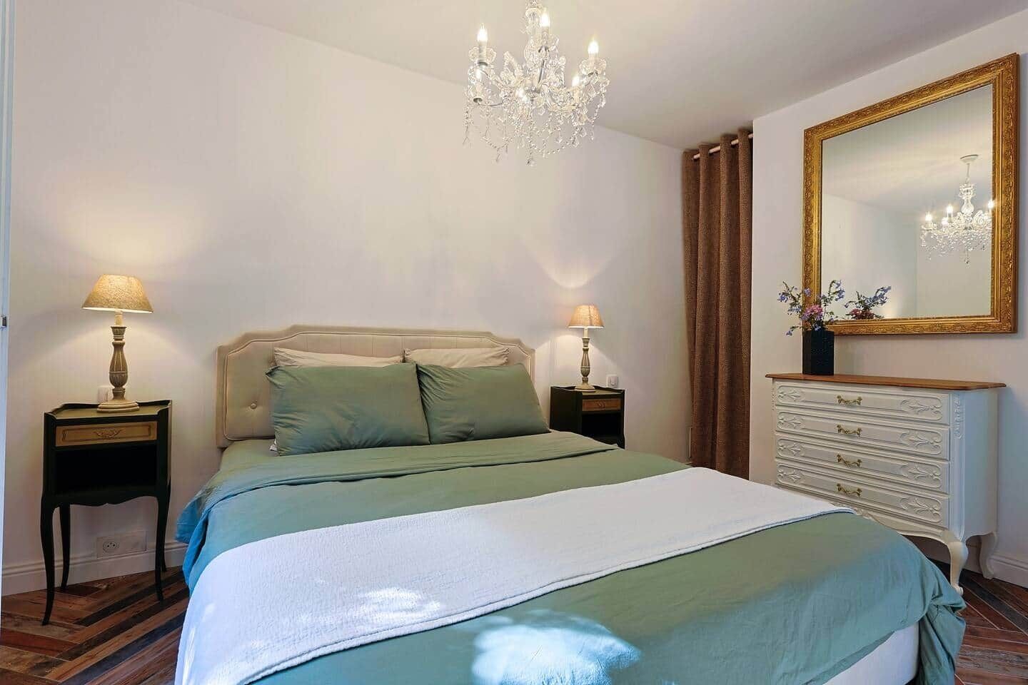 A bedroom with a bed, two bedside tables with lamps, a dresser, a chandelier, and a large mirror.
