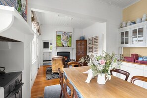 A dining area with a wooden table, chairs, a vase of flowers, and a kitchen in the background. - No 52 Bowral - Sir Donald Bradman`s Childhood Home (Bowral)