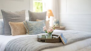 Een net opgemaakt bed met kussens, een nachtkastje met een lamp, en een mand met bloemen en een kaart.