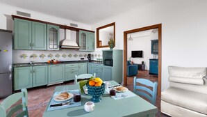 A kitchen with teal cabinets, a dining table with a fruit bowl, and a sofa in the adjacent living area. - Estate4home - Casa Marelia (Sorrento)