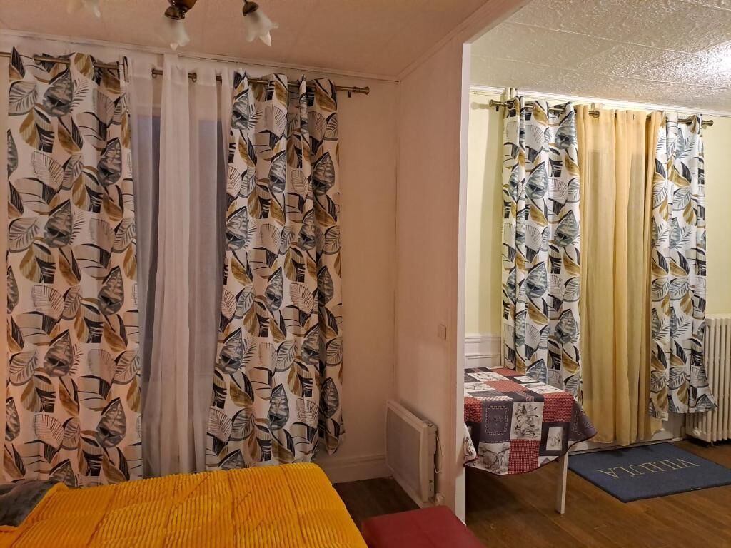 Une pièce comprenant un lit, une petite table recouverte d’une courtepointe, des rideaux à motifs de feuilles et un radiateur.