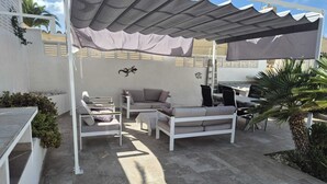 Una terrazza esterna con area salotto coperta, un divano, un tavolo e un piccolo elemento decorativo a parete.