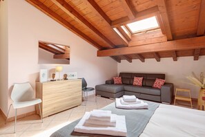 A modern bedroom with a loft-style ceiling, a gray sofa, a wooden dresser, a chair, and a bed with towels.
