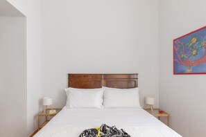Hotellihuone, jossa on puinen sängynpääty, valkoiset vuodevaatteet, kaksi yöpöytälamppua, pieni pöytä lampun kanssa ja värikäs taulu seinällä.
