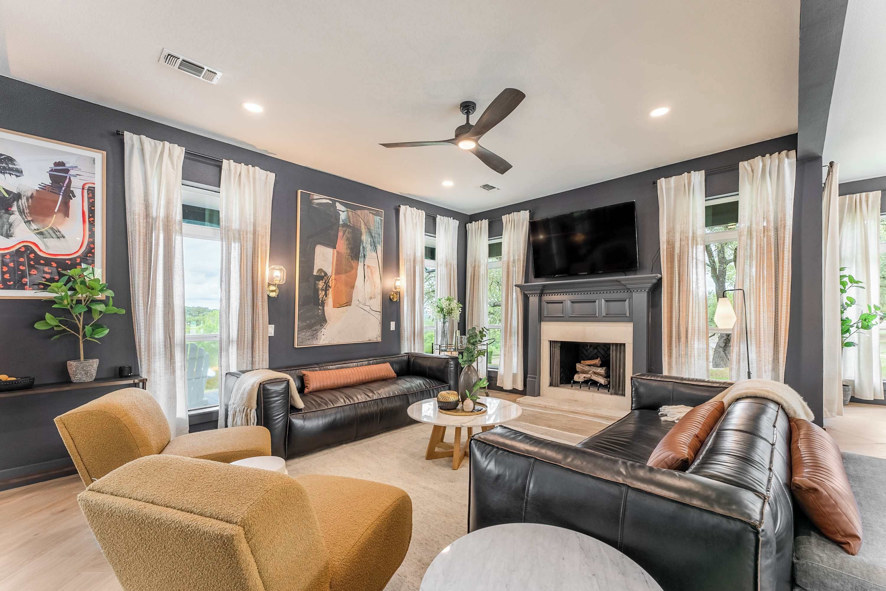 A modern living room with a fireplace, leather sofas, a ceiling fan, and large windows.