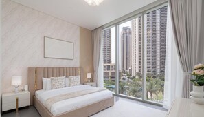 Ein modernes Hotelzimmer mit einem großen Bett, einem Nachttisch, einer Lampe, einem gerahmten Bild an der Wand und einem großen Fenster mit Blick auf die Stadt.