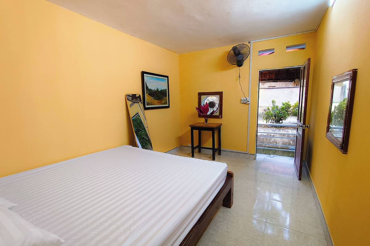 A room with a bed, a fan, and two framed pictures on the wall.