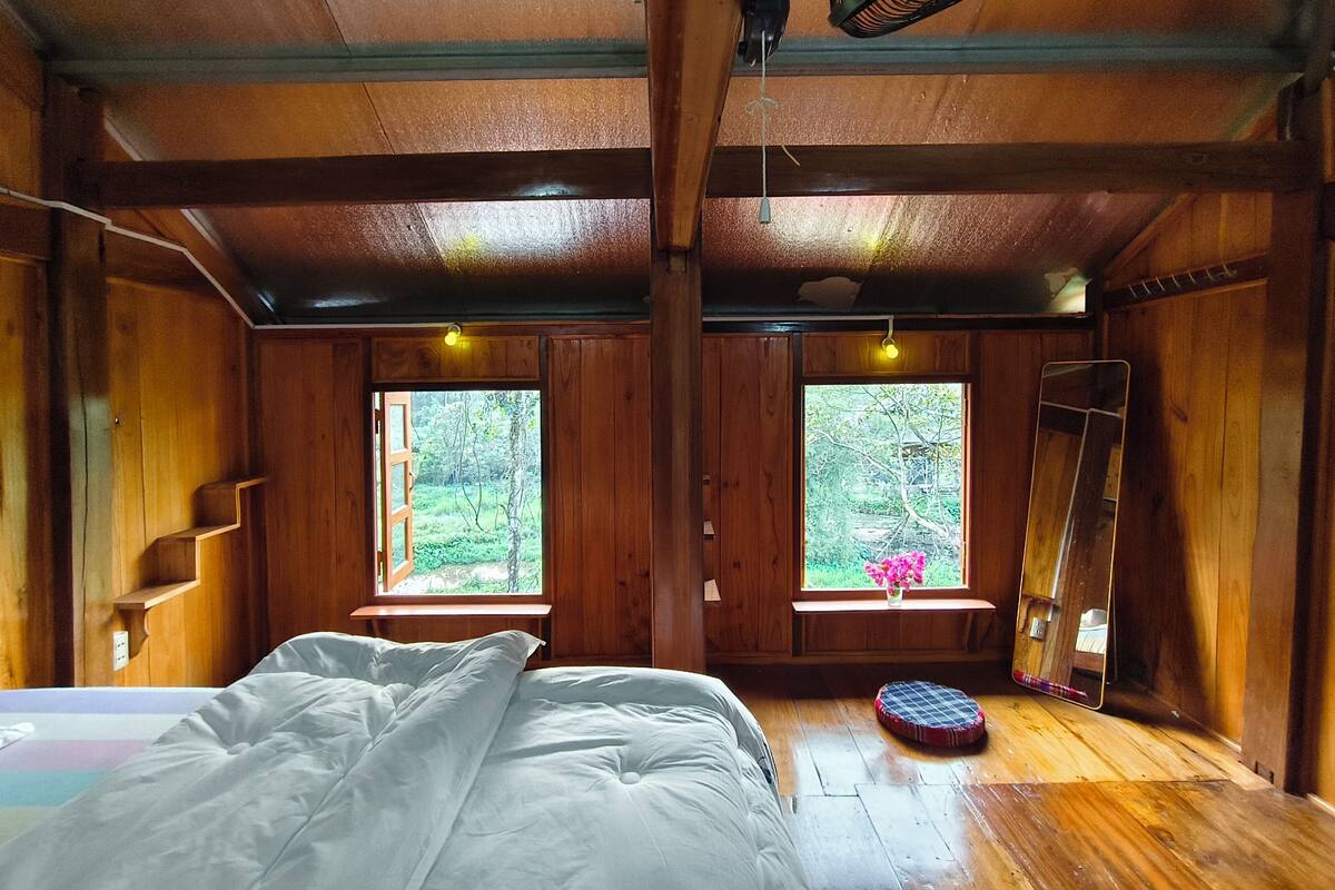 A wooden cabin bedroom with a bed, two windows, a ladder, and a mirror.