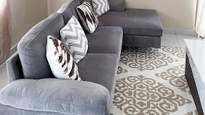 Een moderne woonkamer met een grijze modulaire bank, een getextureerd vloerkleed, wandplanken met decoratieve objecten, en een potplant.