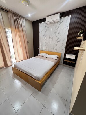 A modern bedroom with a wooden bed, marble-patterned wall, air conditioning unit, and a radiator. - Baski stays  (Serrekunda)