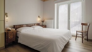 A bedroom with a wooden ceiling, a bed with white bedding, a nightstand with a lamp, a chair, and a window with blinds. - Historic 1940s Watermill: A Designer Retreat in the Italian Alps (Cerete Basso)