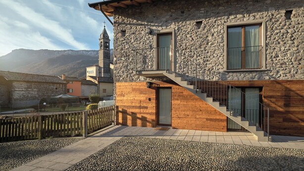 A stone building with a wooden staircase and a view of a mountain and a church. - Historic 1940s Watermill: A Designer Retreat in the Italian Alps (Cerete Basso)