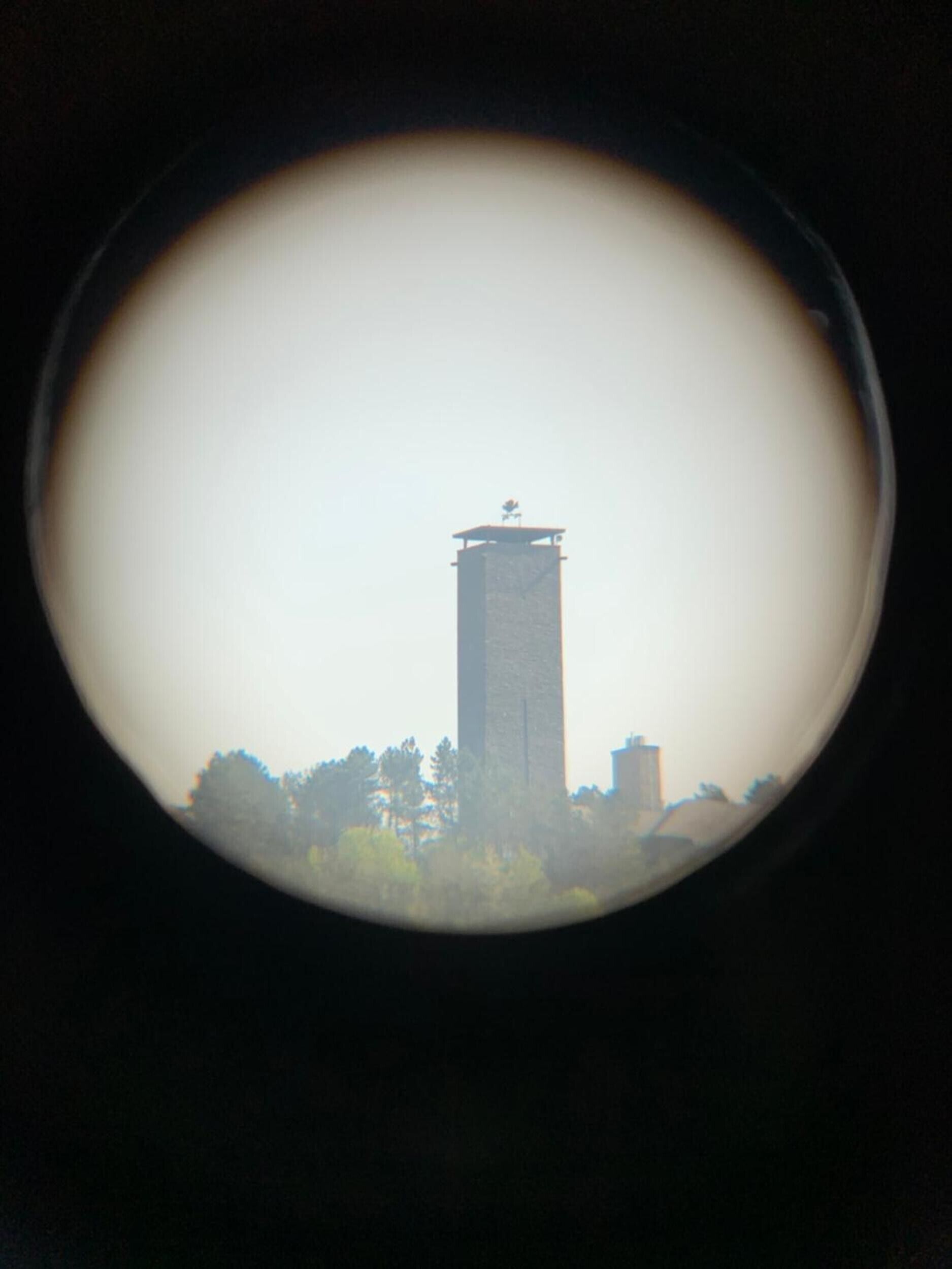 A circular view of a tall, narrow tower with a flat top, surrounded by trees and buildings.