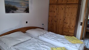 A bedroom with a wooden headboard, a bed with white bedding, a wooden wardrobe, and a painting of a beach scene on the wall. - Holiday Apartment for 4 Guests (64 m²) on Fehmarn (Fehmarn)
