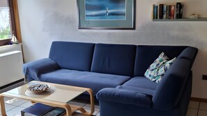 A modern living room with a blue sofa, a glass coffee table, a framed picture above the sofa, and a bookshelf with books. - Holiday Apartment for 4 Guests (64 m²) on Fehmarn (Fehmarn)