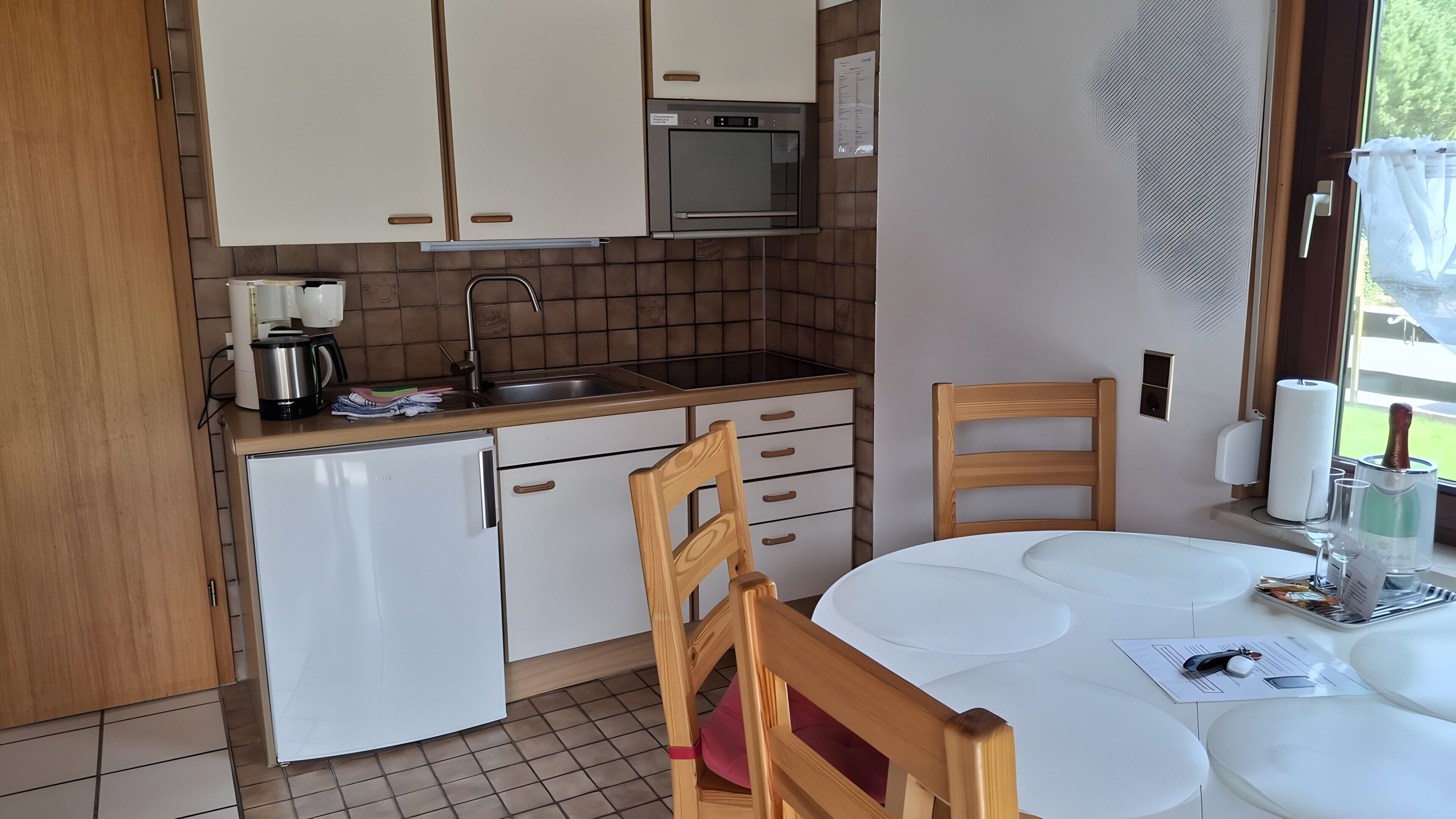 A compact kitchen with a built-in oven, a sink, and a small refrigerator. There's a dining table with chairs, and a window with a view of greenery outside.