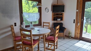 A room with a round table, four wooden chairs with red cushions, a clock on the wall, a television on a wooden shelf, and a window with a view of greenery. - Holiday Apartment for 4 Guests (64 m²) on Fehmarn (Fehmarn)