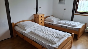 Twee houten stapelbedden met wit beddengoed, een houten dressoir met een lamp, en een raam met uitzicht op groen.