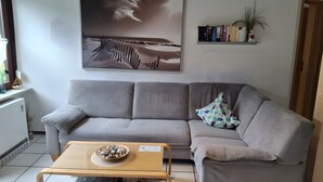 Een woonkamer met een grijze bank, een houten salontafel met een schaal stenen, een boekenkast met boeken, een aan de muur bevestigde plant en een schilderij van een strandgezicht.