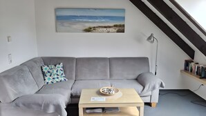 Ein graues Sofa mit einem gemusterten Kissen, ein kleiner Holztisch mit einer Schale, ein Gemälde einer Strandansicht und eine Treppe mit schwarzem Geländer.