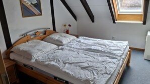Ein Schlafzimmer mit einem hölzernen Bett, weißer Bettwäsche, einem kleinen Nachttisch, einem Wandbild und einem Fenster mit Blick auf Bäume.