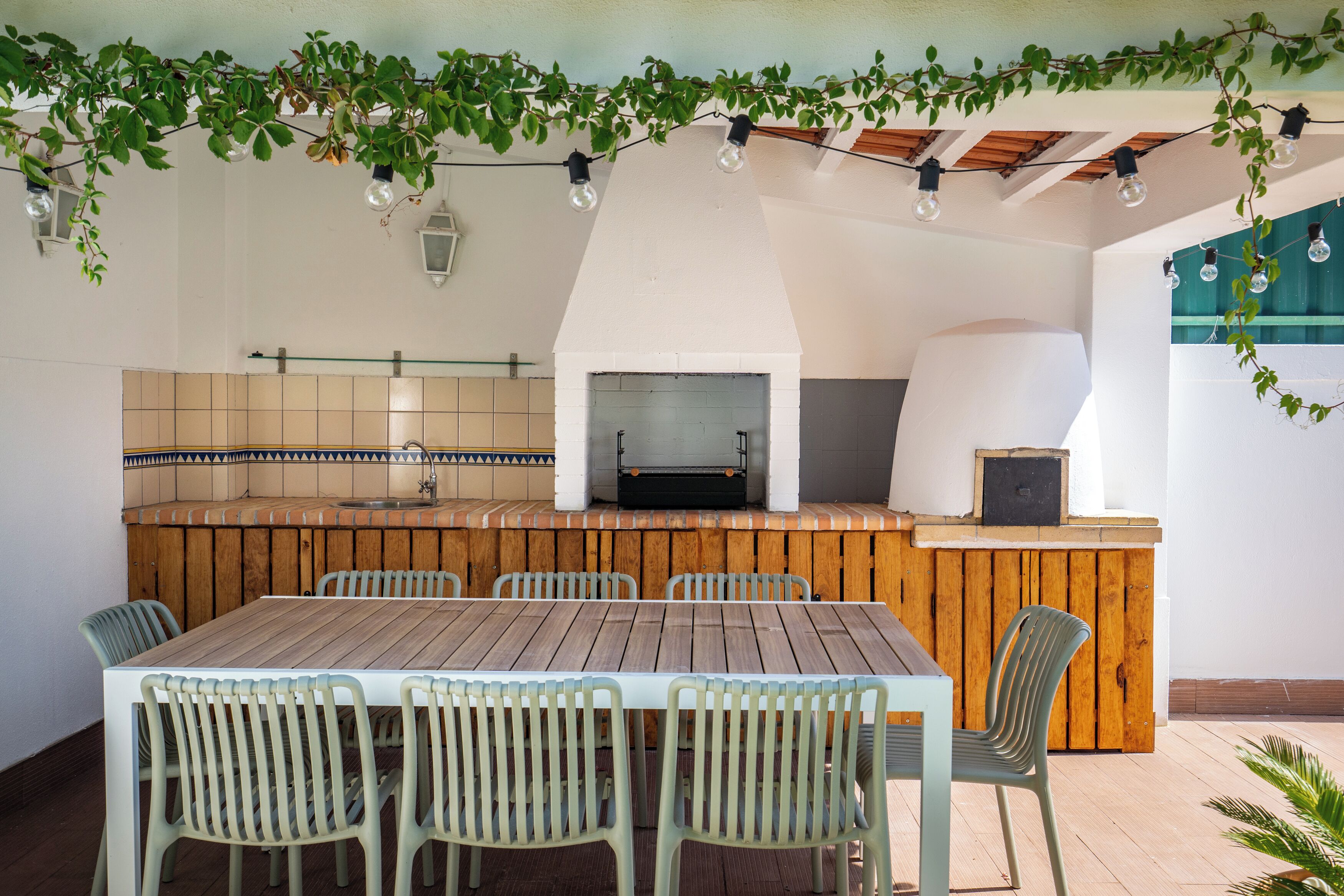 Ein Essbereich im Freien mit Holztisch und -stühlen, einem Backofen aus Ziegelsteinen und einer gefliesten Spritzschutzwand.