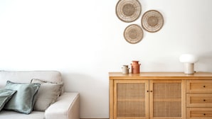 Una habitación con un sofá de color claro, un mueble de madera con cajones y tres adornos de pared tejidos.