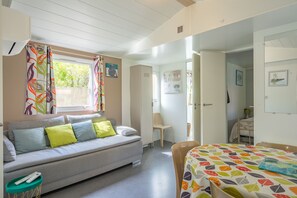 A room with a sofa, a bed, a chair, a small table, and a window with curtains. - Mobile Home near Royan-Oléron with Shared Pool, Private Terrace & Air Conditioning (Saint-Sornin)