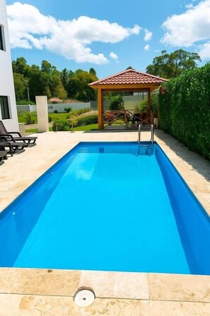 Una piscina rectangular con solárium empedrado, un pequeño pabellón y un edificio blanco al fondo.
