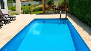 Una piscina rectangular amb una terrassa enrajolada, un petit pavelló i un edifici blanc al fons.